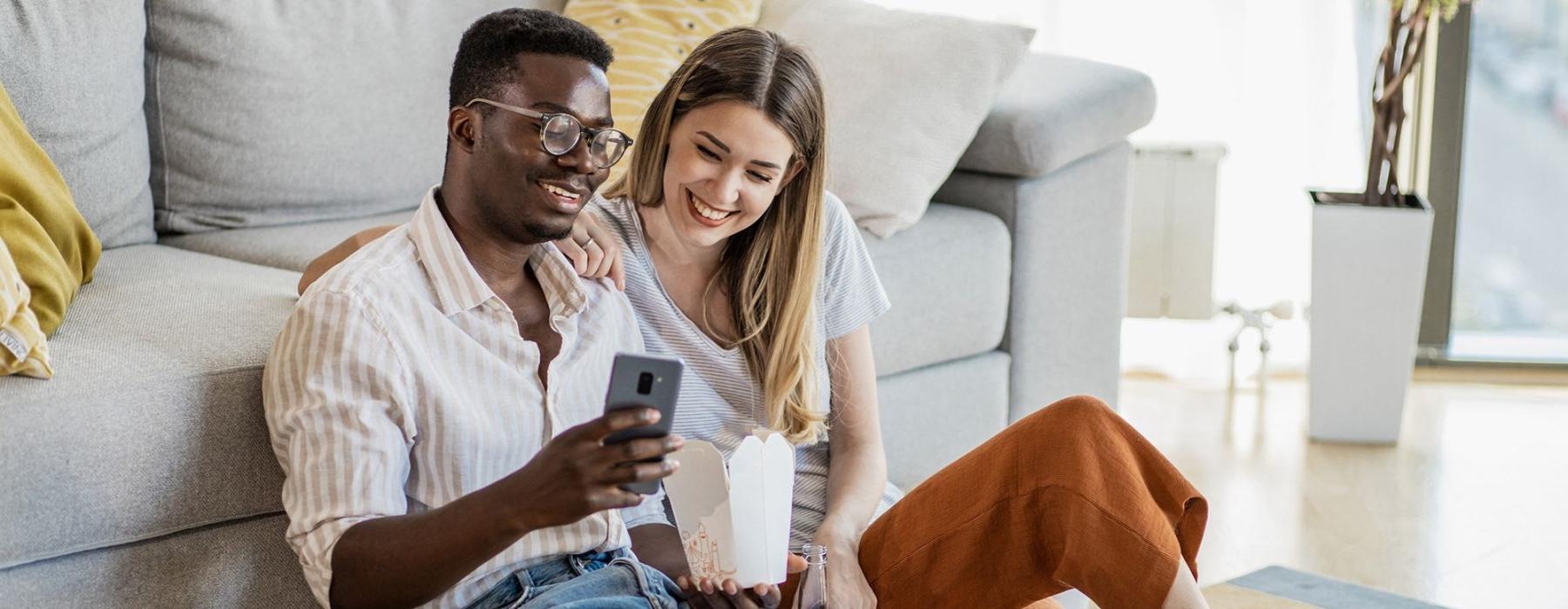 a man and woman with take out, sit against a couch on their living room floor and watch at their cell phone